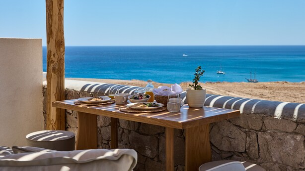 A wooden table with breakfast items and a potted plant, overlooking a beach with boats and clear blue water. - Armony Suites Milos (Milos)