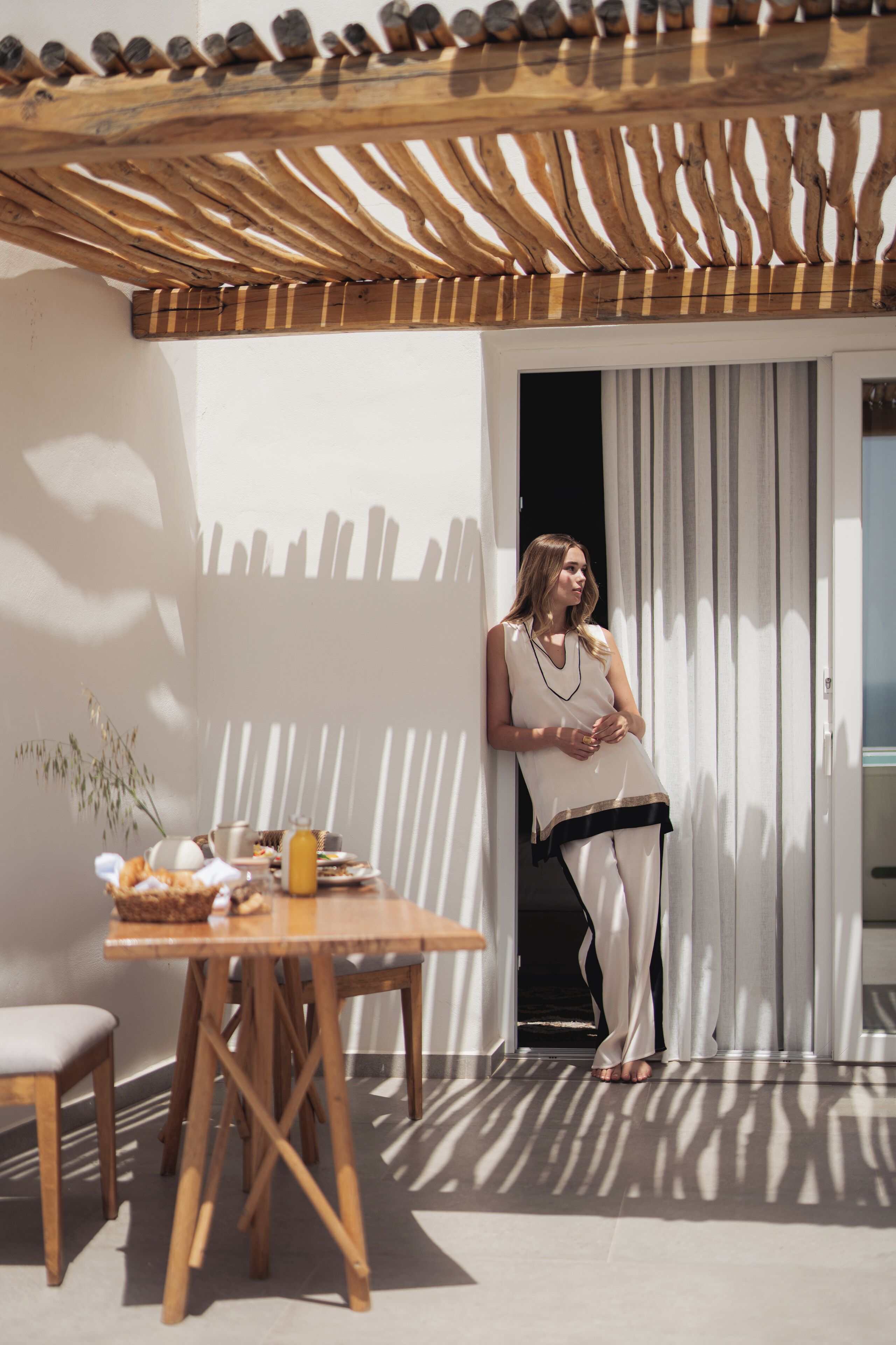 Una persona in piedi accanto a un tavolo con cibo e bevande, una pergola in legno e un muro bianco.