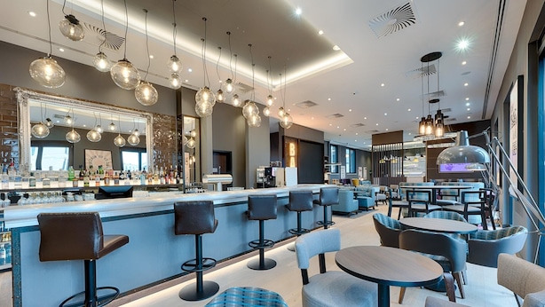 A modern bar area with a long counter, multiple bar stools, and round tables. - Premier Inn Regensburg City Centre (Regensburg)