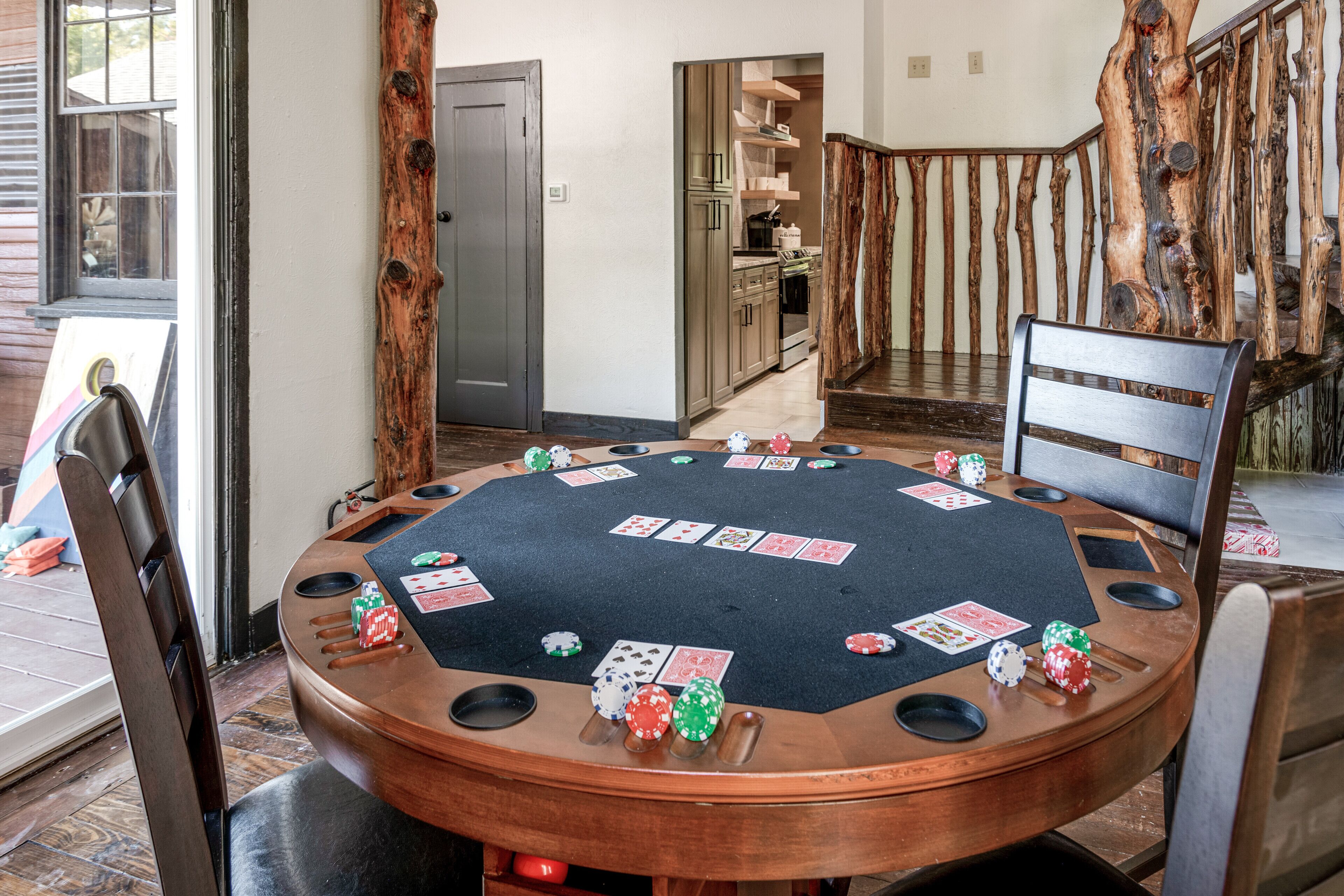 A poker table with chips and cards, surrounded by wooden chairs, in a room with a rustic staircase and a kitchen in the background.