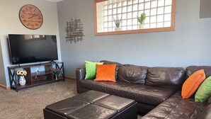 A living room with a sectional sofa, a flat-screen TV, a wooden clock, and a window with a plant. - Private Downtown Apt, King Bed, Jet Bath, Fireplace, Grill, near Indiana Dunes (Chesterton)
