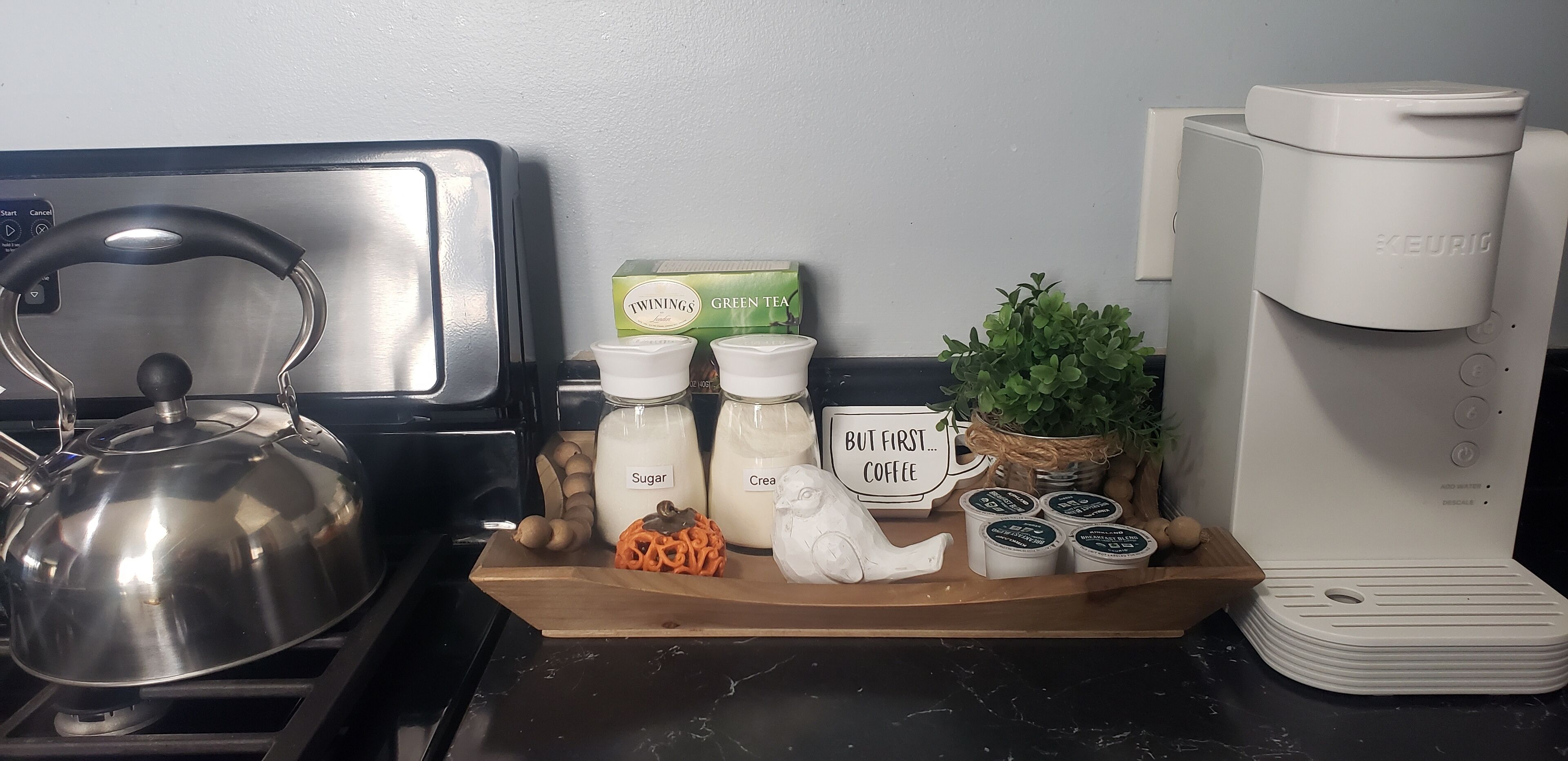 A kitchen counter with a kettle, sugar and creamer containers, Twinings green tea box, a coffee maker, and a small plant.