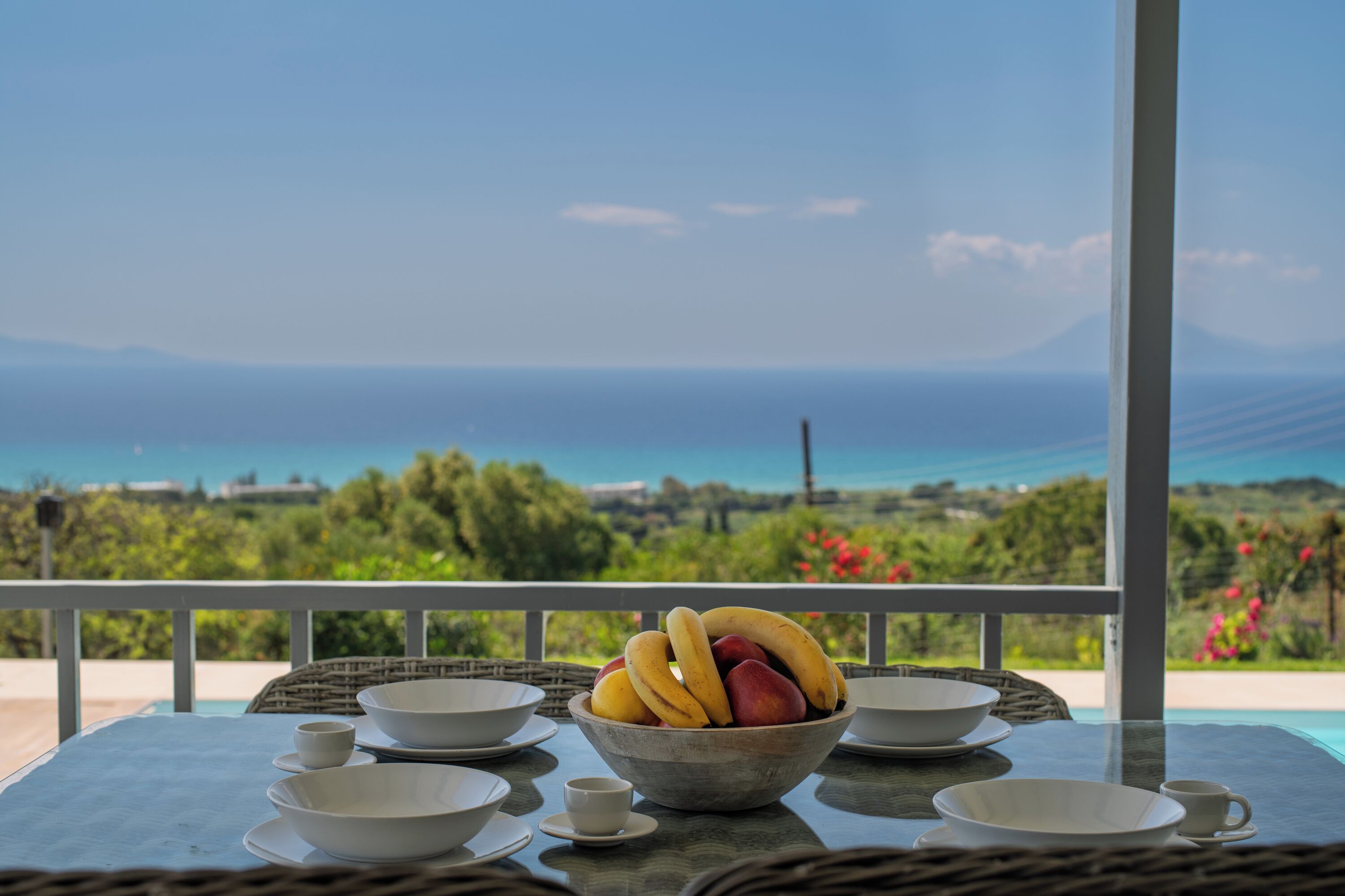 A table with a bowl of fruit, white bowls, and a view of the sea and greenery.