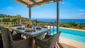Un espacio de estar techado al aire libre con una mesa lista para comer, con vistas a una piscina y un paisaje marino y montañoso.
