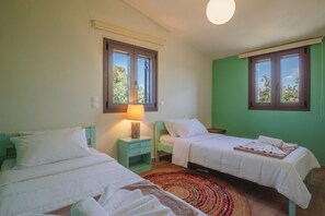 A room with two beds, a nightstand with a lamp, a woven rug, and large windows with a view of trees.
