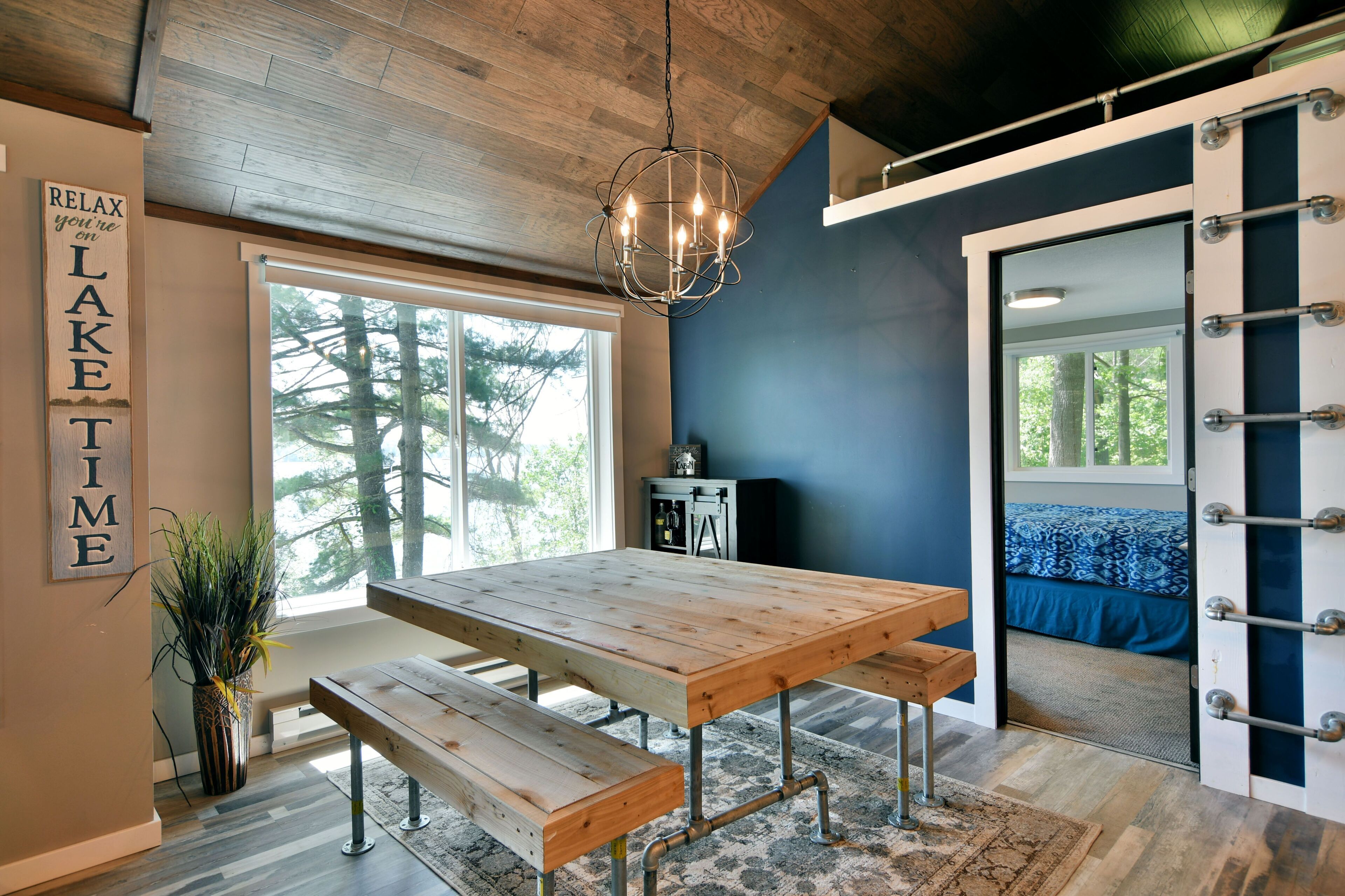 Un interior moderno con una mesa de comedor de madera, bancos, una lámpara colgante y vista a un bosque a través de un ventanal grande.