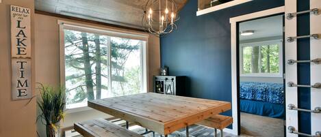 Un interior moderno con una mesa de comedor de madera, bancos, una lámpara colgante y vista a un bosque a través de un ventanal grande.