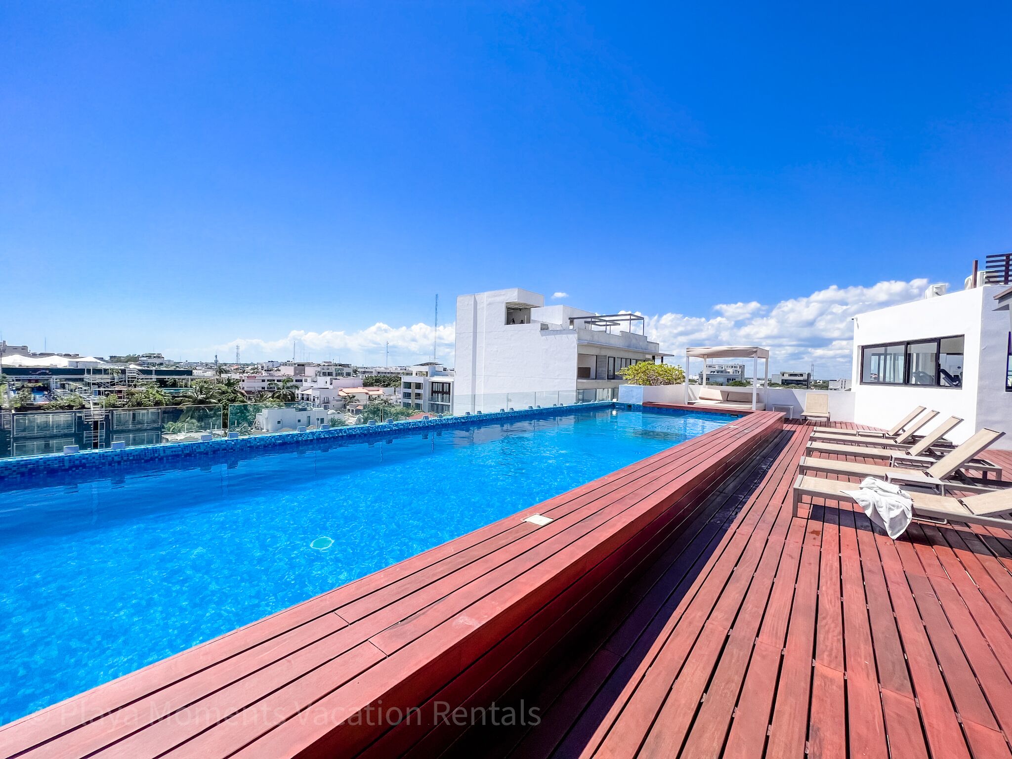 A rooftop pool with wooden deck and lounge chairs, surrounded by modern buildings and a clear sky.
