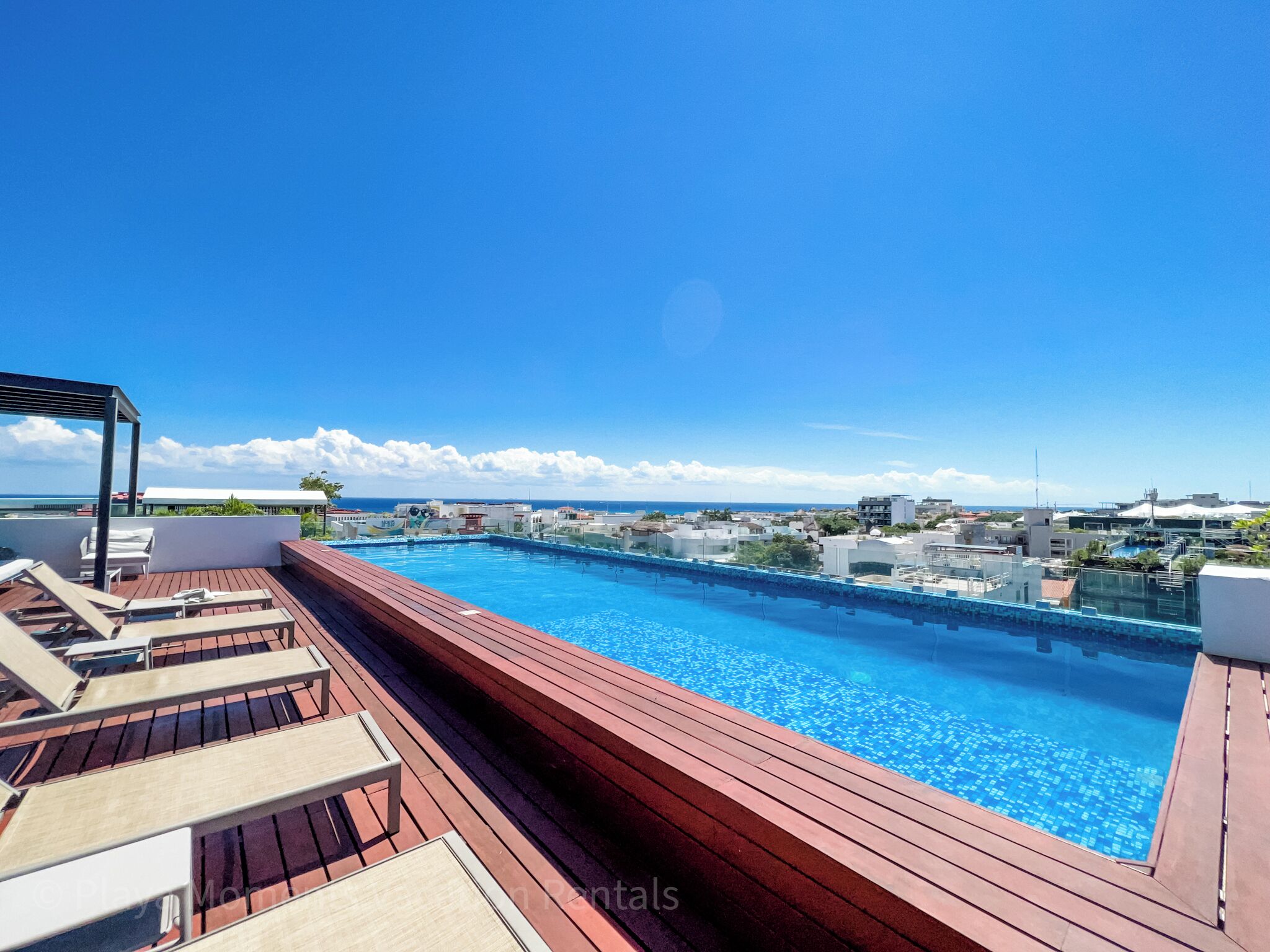 A rooftop pool with wooden deck and lounge chairs, overlooking a cityscape and ocean.