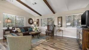 A spacious living room with a large brown leather sofa, a green armchair, and a wooden coffee table. There is a television on a wooden cabinet, and the room has large windows with blinds. - Treehouse Cottage | 2 Bed, 1 Bath (Canyon Lake)