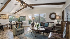 A spacious living room with a high ceiling, a ceiling fan, and a large window. There is a brown leather sofa, a green armchair, and a coffee table with a vase of flowers. - Treehouse Cottage | 2 Bed, 1 Bath (Canyon Lake)