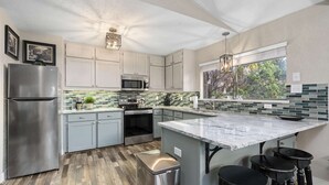 A modern kitchen with grey cabinets, stainless steel appliances, a large island with bar stools, and a view through the window. - Treehouse Cottage | 2 Bed, 1 Bath (Canyon Lake)