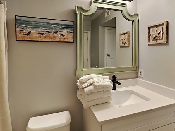 A bathroom with a white sink, a green-framed mirror, a painting of birds on the wall, and a stack of white towels.