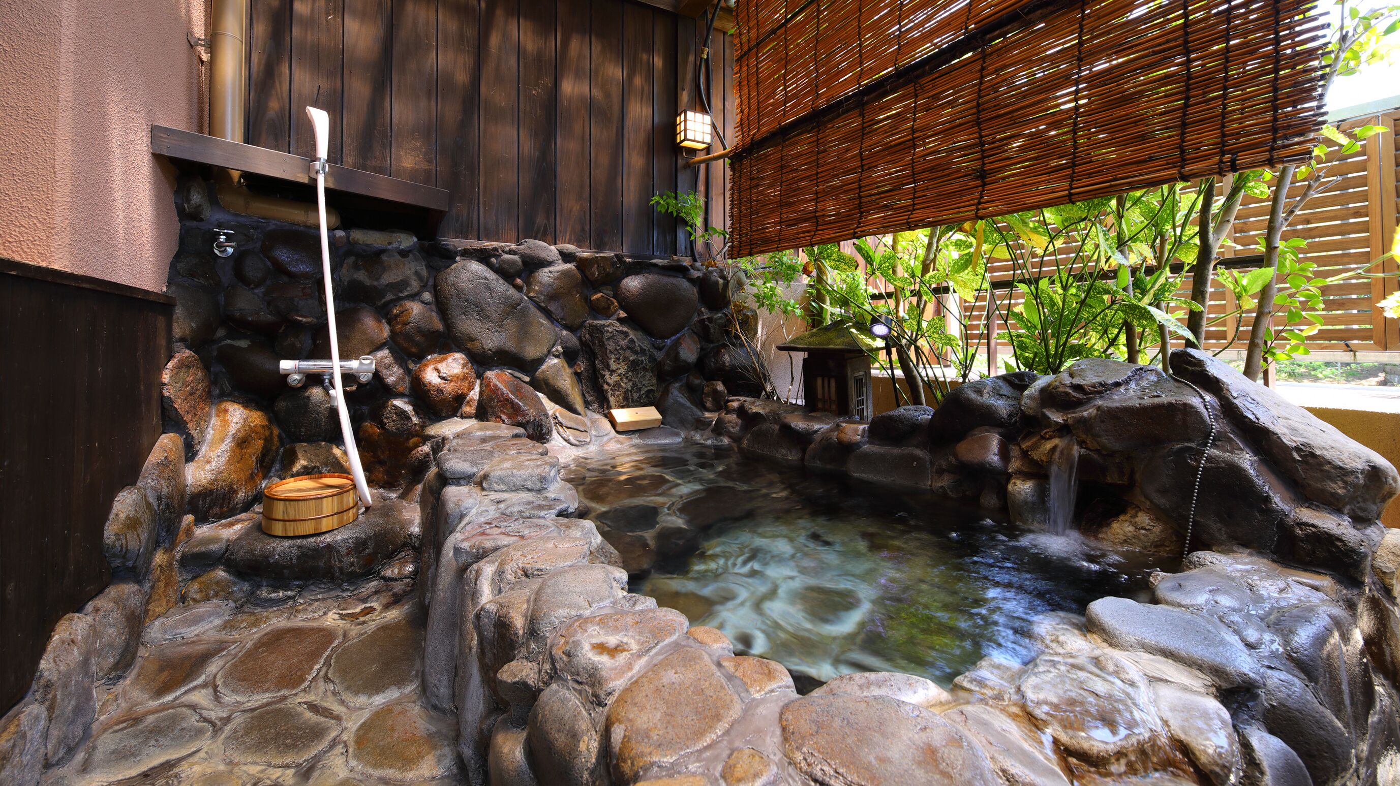 A stone hot spring with a wooden structure, a stone wall, and a bamboo fence.