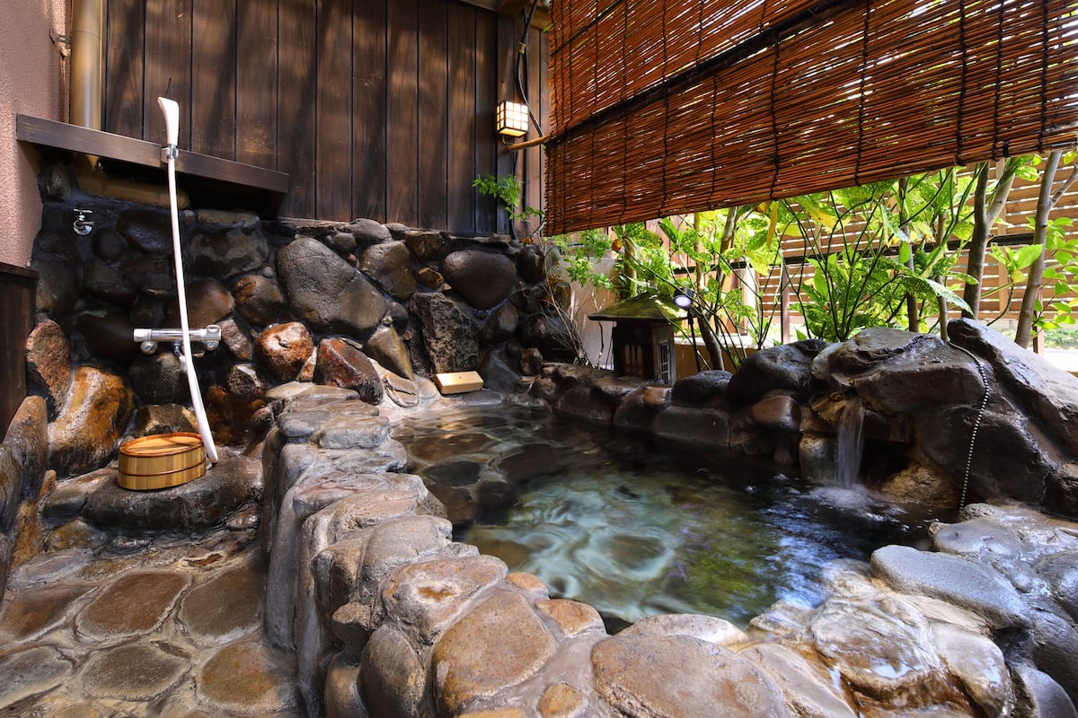 A stone hot spring with a wooden structure, a stone wall, and a bamboo fence.