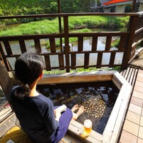 A person sitting by a wooden pool with a drink, overlooking a river and greenery.