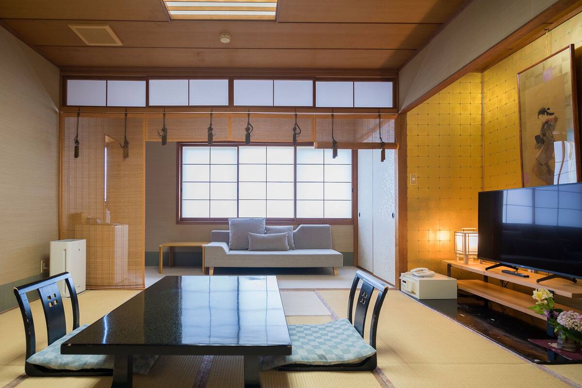 A traditional Japanese-style room with tatami flooring, a low table, cushions, a sofa, a television, and a sliding door.