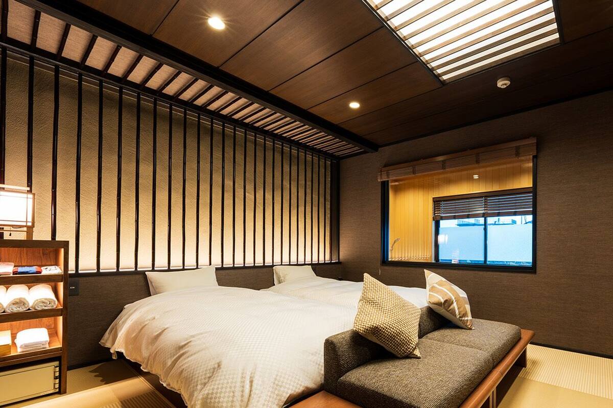 A hotel room with a bed, a sofa, a window with blinds, and a shelf with towels.