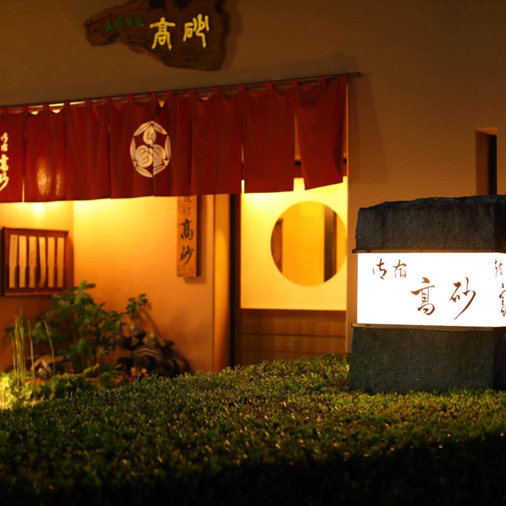 Una entrada tradicional japonesa con toldo rojo, un letrero con caracteres japoneses y un pequeño jardín delantero.
