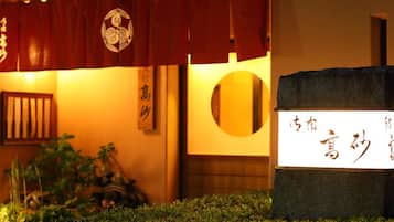 Una entrada tradicional japonesa con toldo rojo, un letrero con caracteres japoneses y un pequeño jardín delantero.