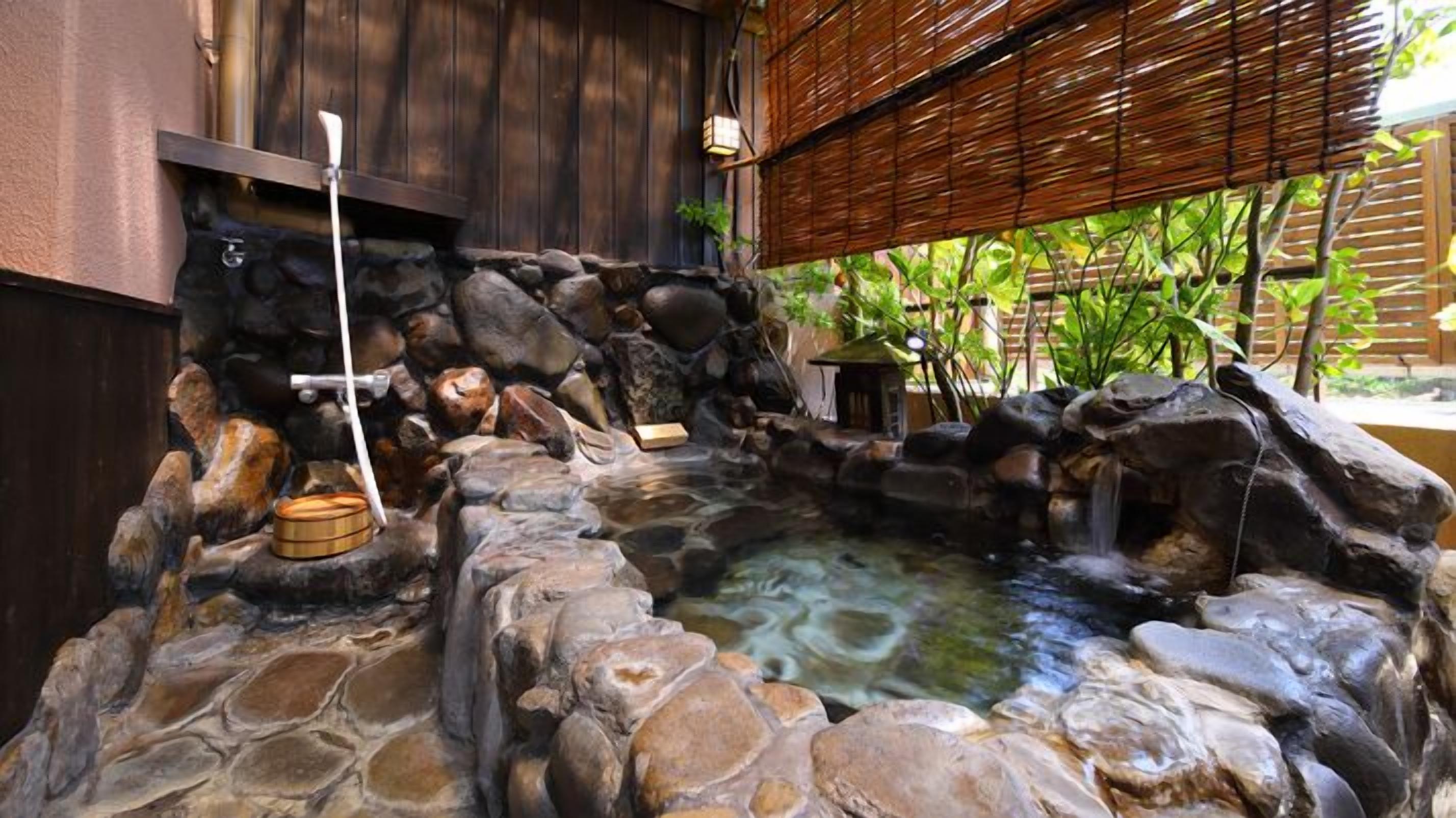 A stone hot spring bath area with a wooden structure, a stone basin, and a small waterfall.