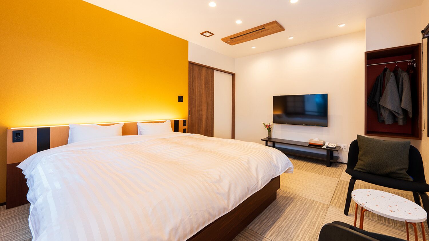 A hotel room with a large bed, a TV mounted on the wall, a seating area with a chair and table, and a wardrobe with hanging clothes.