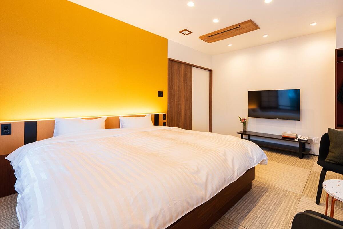A hotel room with a large bed, a TV mounted on the wall, a seating area with a chair and table, and a wardrobe with hanging clothes.