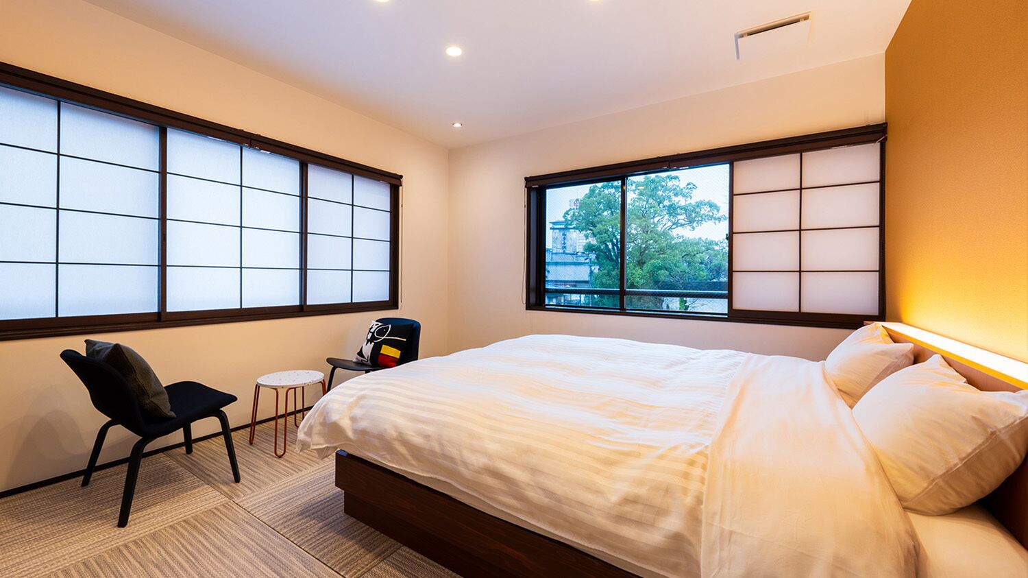 A bedroom with a bed, a chair, a small table, and large windows with a view.