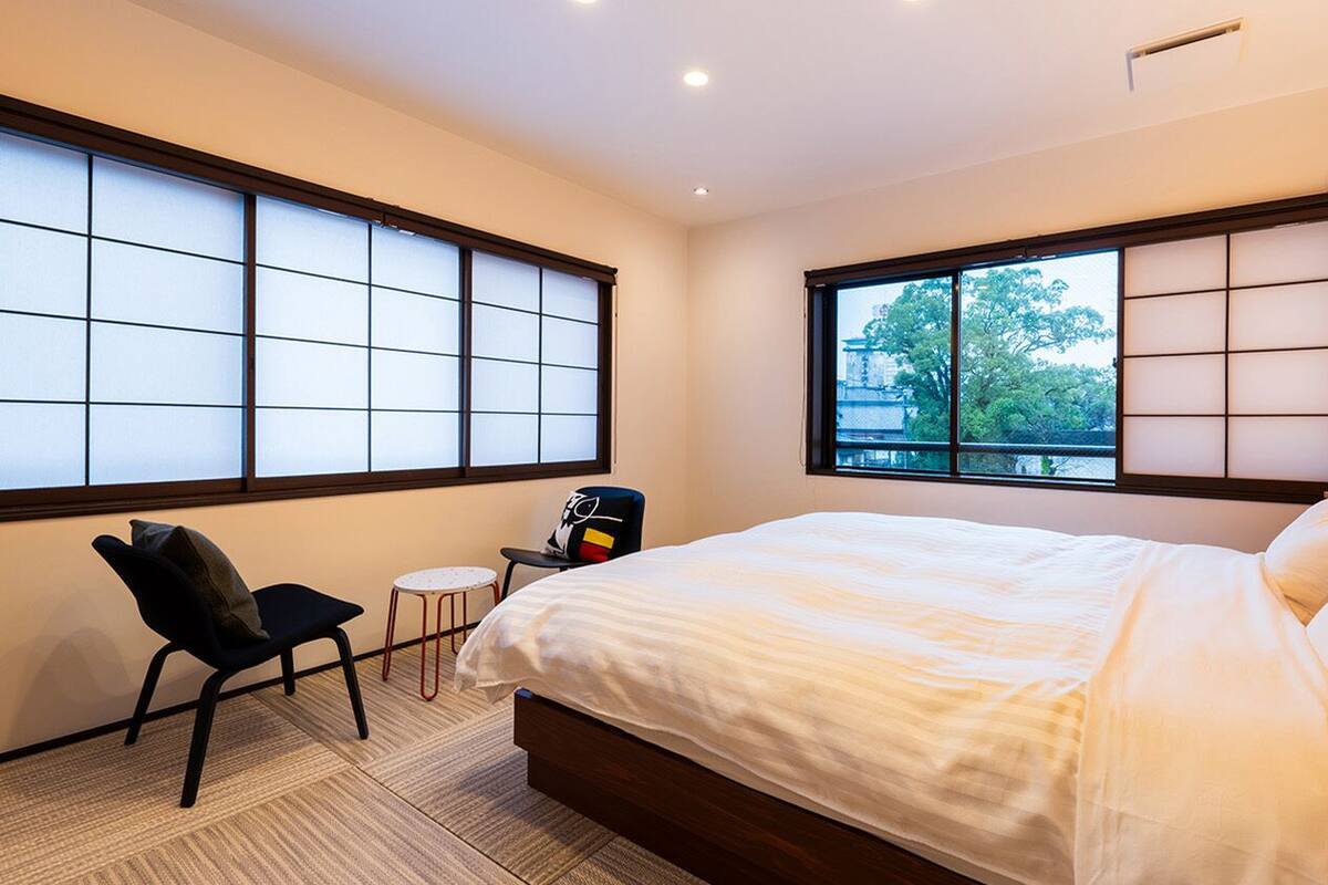 A bedroom with a bed, a chair, a small table, and large windows with a view.