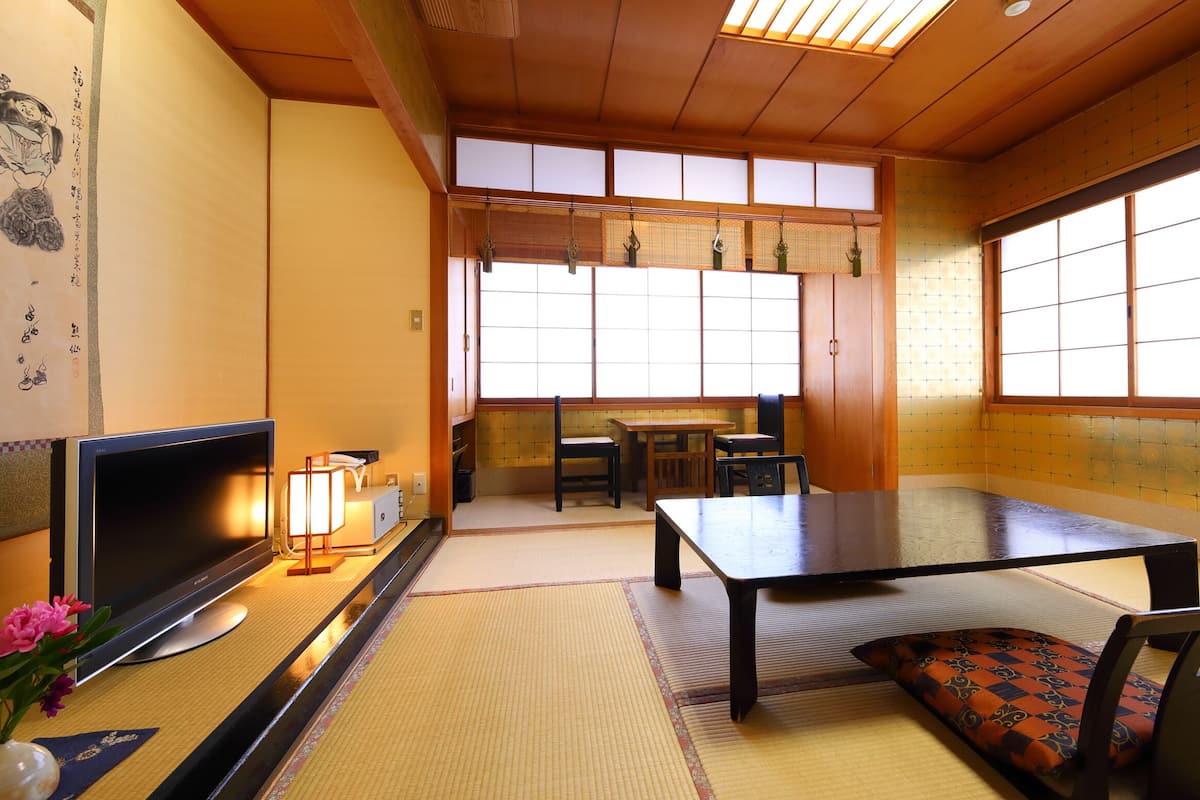A traditional Japanese room with tatami flooring, a low table, chairs, a television, and a window with shoji screens.