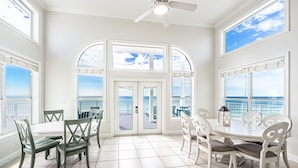 A spacious dining area with a view of the ocean, featuring a white table and chairs, and a ceiling fan.