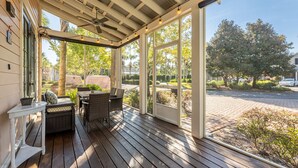 A covered outdoor patio with wooden flooring, wicker furniture, and large windows offering a view of the street and greenery.