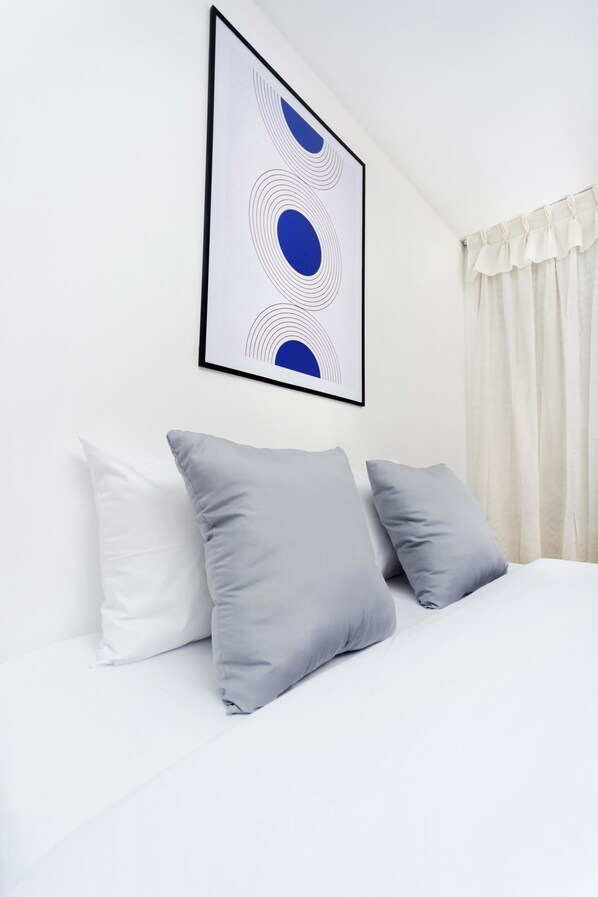 Un lit bien fait, avec des oreillers blancs et gris, une œuvre d’art abstraite encadrée au-dessus, et un rideau blanc sur le côté droit.