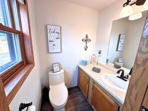 A bathroom with a white toilet, wooden cabinets, a sink, and a mirror. - Edgewater Retreat on Beautiful Soo Lake! (Phillips)