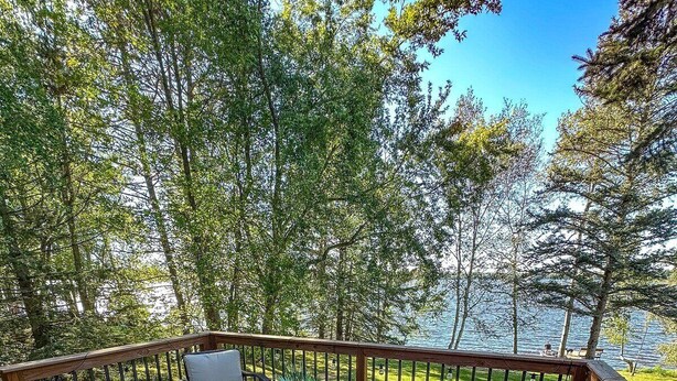 A wooden deck with wicker furniture, including a sofa and chairs, surrounded by lush greenery and a view of a body of water. - Edgewater Retreat on Beautiful Soo Lake! (Phillips)