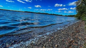 A rocky shoreline with clear blue water and a distant tree line under a partly cloudy sky. - Edgewater Retreat on Beautiful Soo Lake! (Phillips)