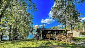 A rustic cabin with a wooden deck and a porch, surrounded by lush greenery and a clear blue sky. - Edgewater Retreat on Beautiful Soo Lake! (Phillips)