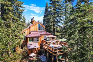 A wooden cabin with a deck and multiple windows, surrounded by dense forest. - Amazing Architecture - Views all around VH#113 by Bear Valley Vacation Rentals (Arnold)
