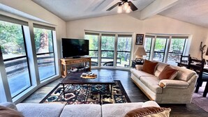 A spacious living room with large windows, a flat-screen TV, a wooden coffee table, and a beige sofa. - In the Pines, Cozy Retreat with Amazing Views (Idyllwild)