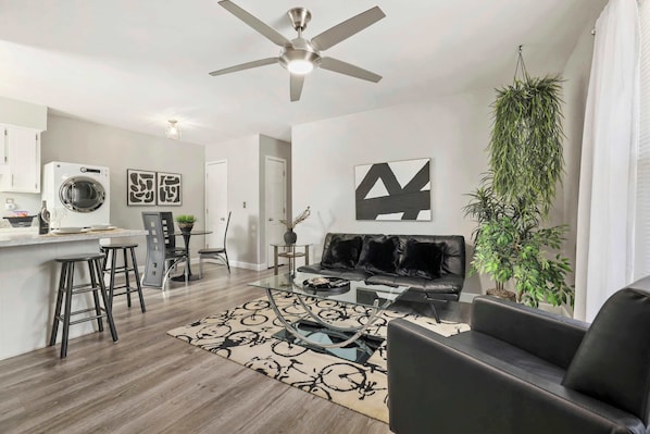A modern living room with a sofa, coffee table, and bar stools. There is a ceiling fan, a washing machine, and a large plant.