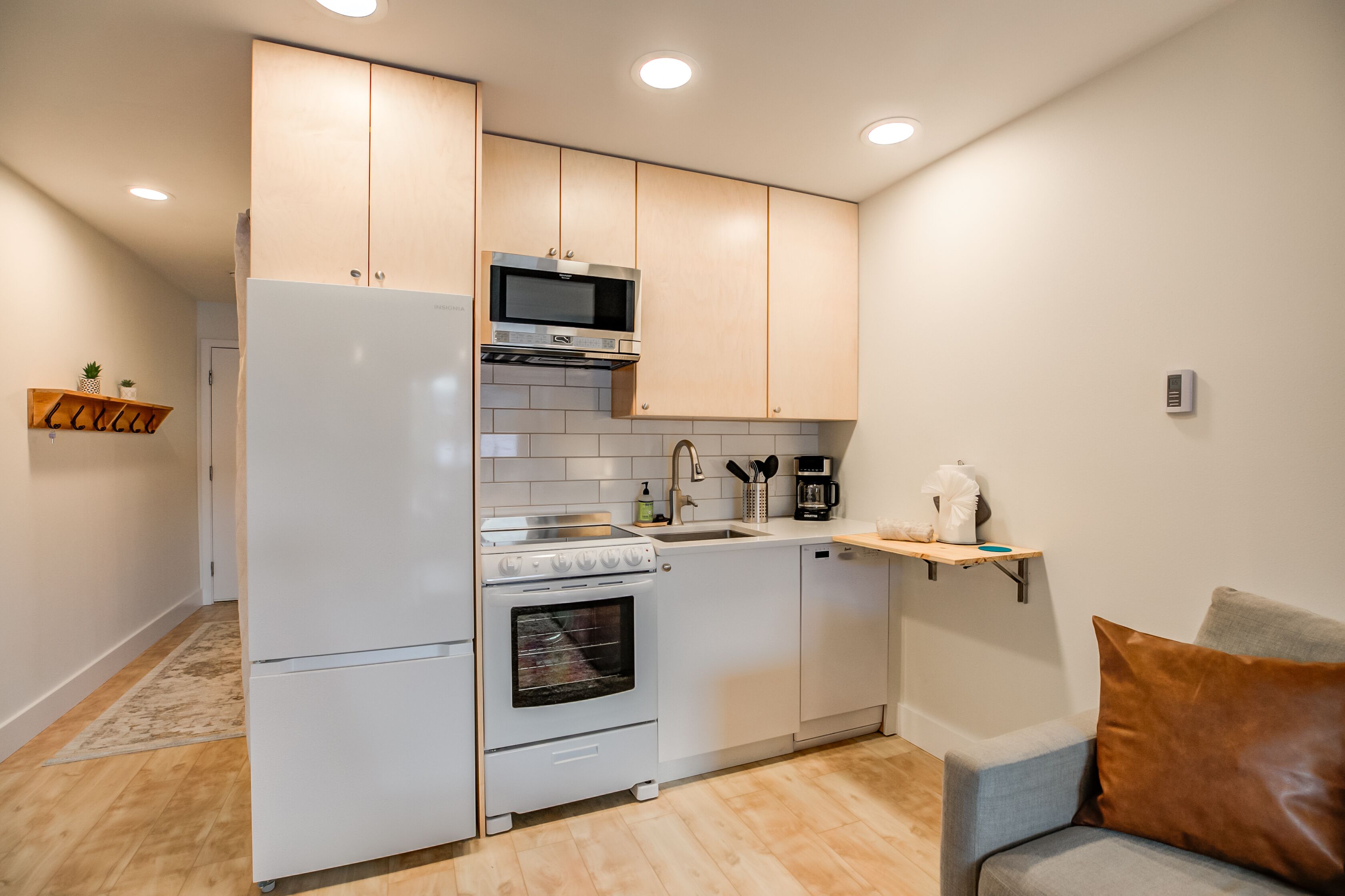 A compact kitchen with white appliances, a microwave, a stove, and a dishwasher. There is a small shelf with kitchen utensils and a coffee maker.