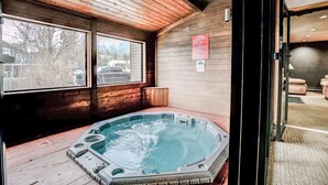 A hot tub in a room with wooden walls and large windows, a wooden ceiling, and a visible door.