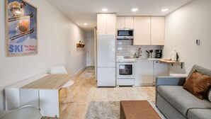 A modern kitchen with white cabinets, a microwave, and a refrigerator. There is a dining area with a wooden table and white chairs. A gray sofa with a brown pillow is in the living area.