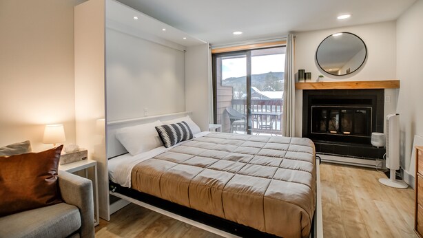 A modern bedroom with a bed, bedside tables, a sofa, a fireplace, and a large window offering a view of the outdoors.