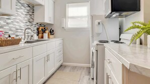 Et moderne køkken med hvide skabe, et flisebeklædt backsplash, en indbygget komfur og et vindue med persienner.