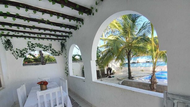 A covered patio with a dining table and chairs, a view of a swimming pool, and a palm tree. - Stay.Plus Nyumba Tulivu Watamu (Watamu)