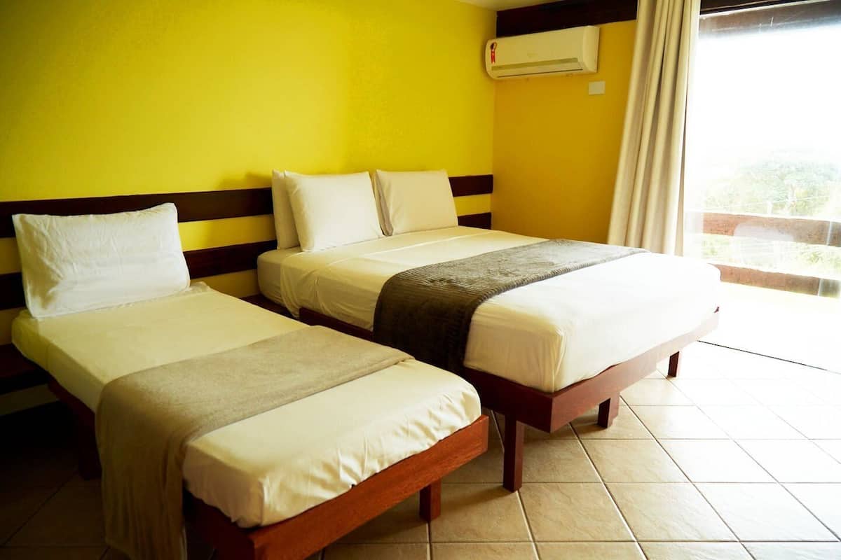 Quarto de hotel com duas camas de solteiro, uma janela com cortinas e um aparelho de ar condicionado na parede.
