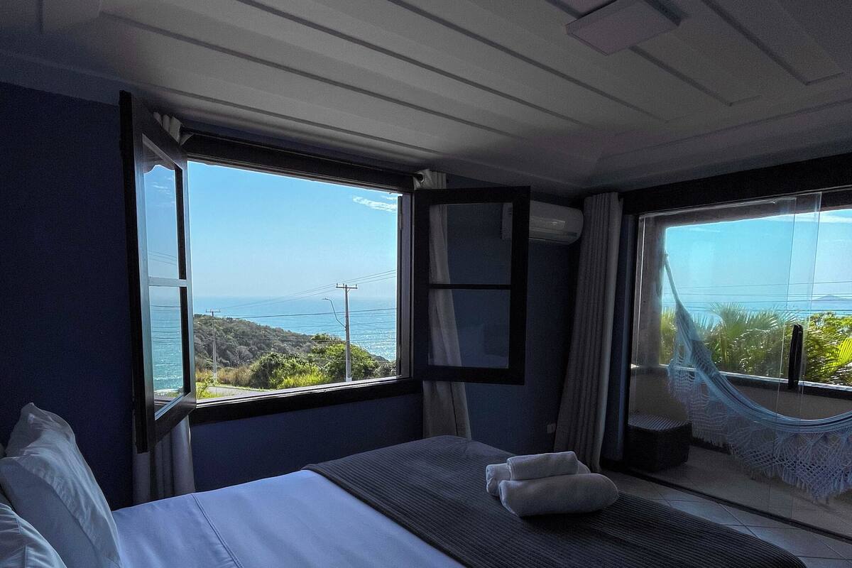 Quarto de hotel com uma cama grande, vista para o mar e uma rede perto da janela.