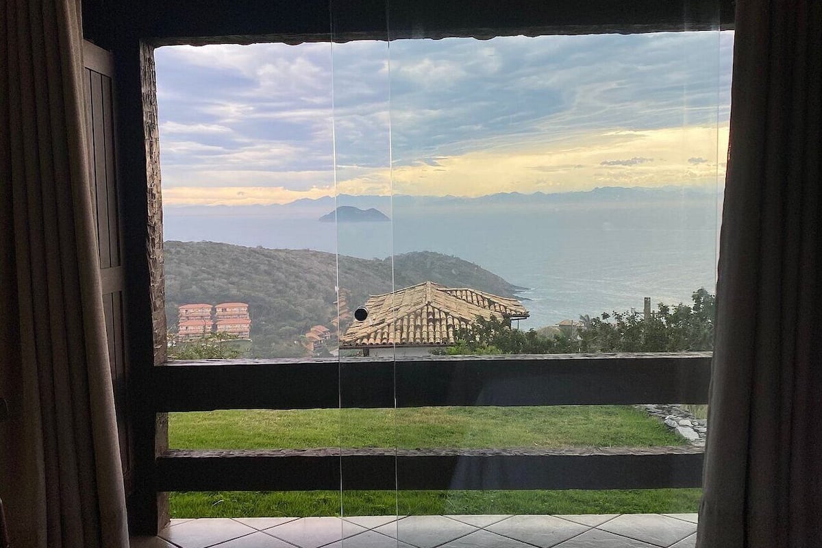 Quarto com uma grande janela que oferece vista para a paisagem costeira, incluindo uma praia e montanhas ao longe.