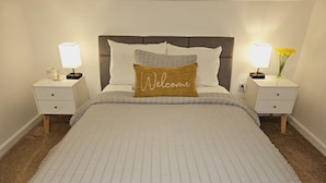 Una cama bien hecha con una almohada que dice 'Welcome', dos mesitas de noche con lámparas y un jarrón con flores en una de ellas.